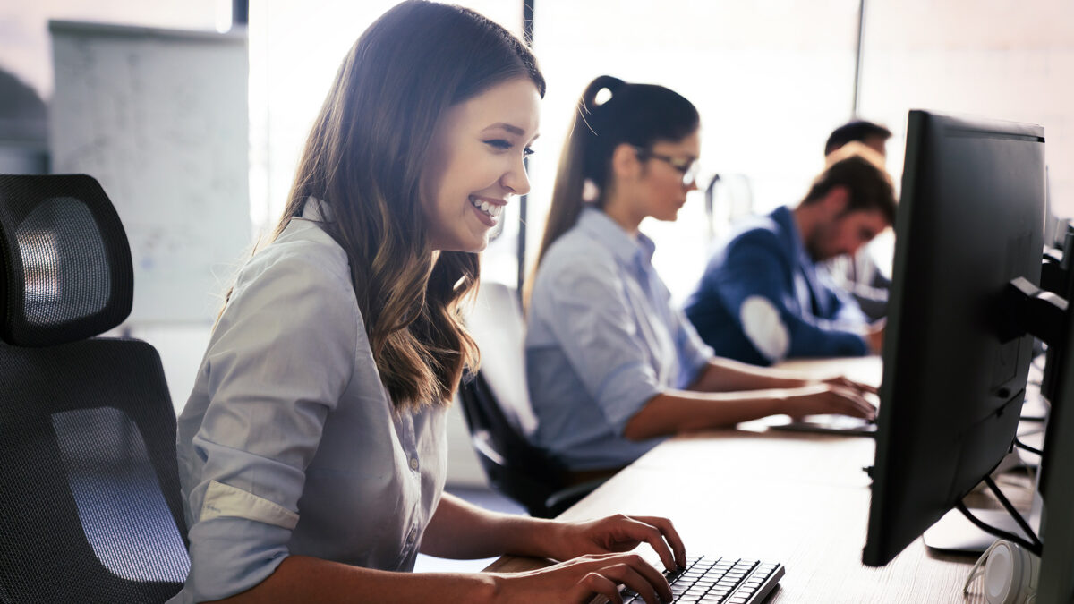 Side view of young woman smiling at computer with coworkers in the background