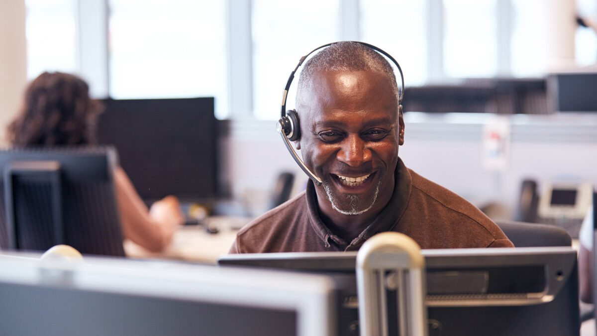 Mature Businessman Wearing Telephone Headset Talking To Caller In Customer Services Department