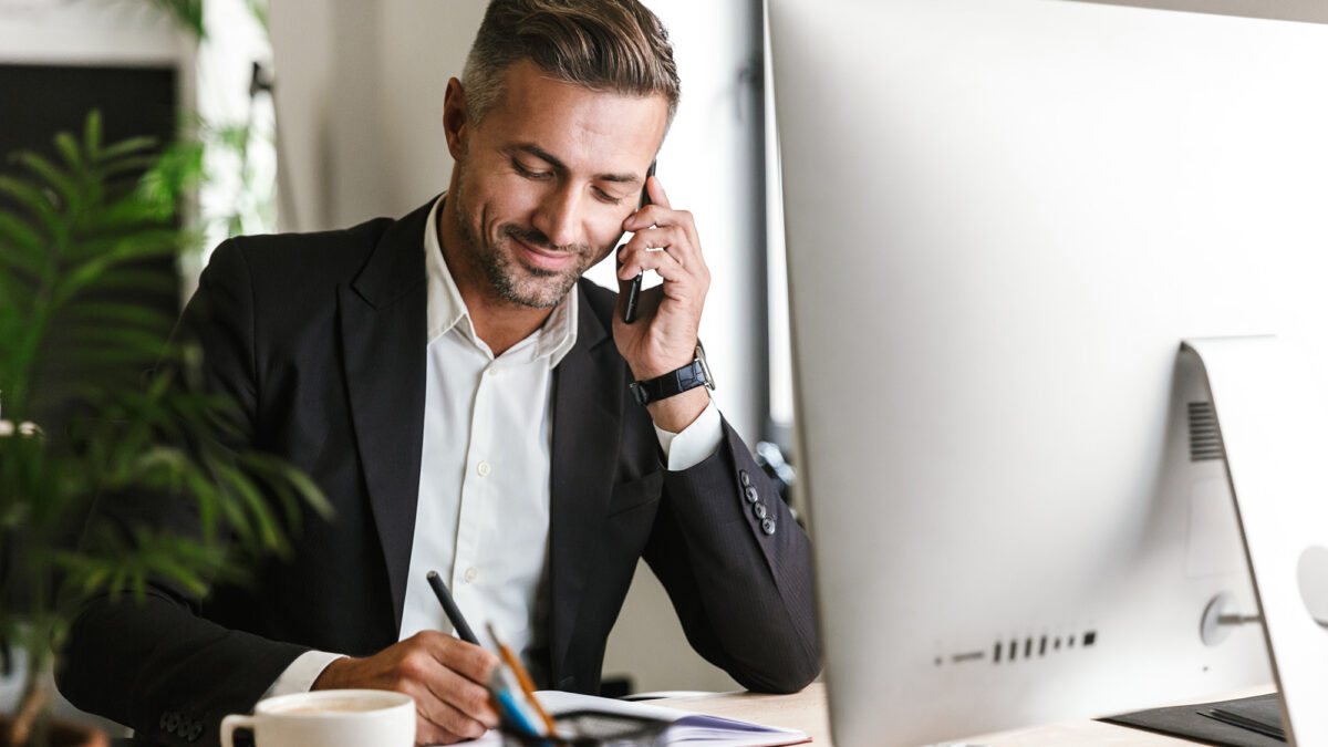 Image of handsome businessman talking on cell phone while working on computer in office