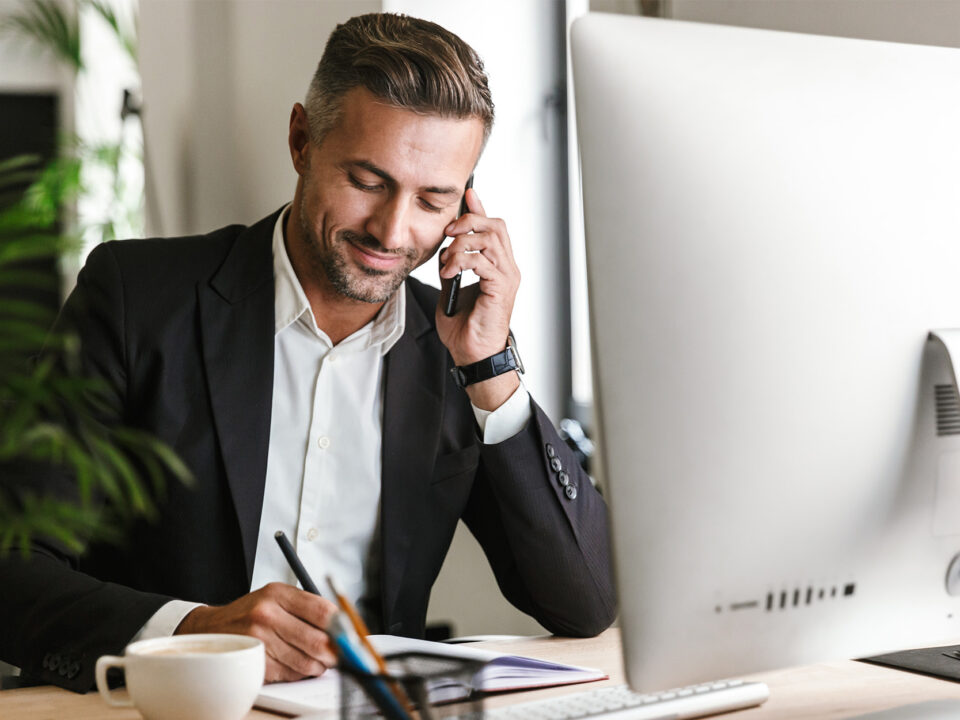 Image of handsome businessman talking on cell phone while working on computer in office